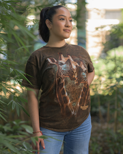 A person standing outdoors. They are wearing a brown t-shirt that features an image of two giraffes with the word “Rainforest Cafe” written below. The setting appears to be a lush green area, likely a garden or park. 