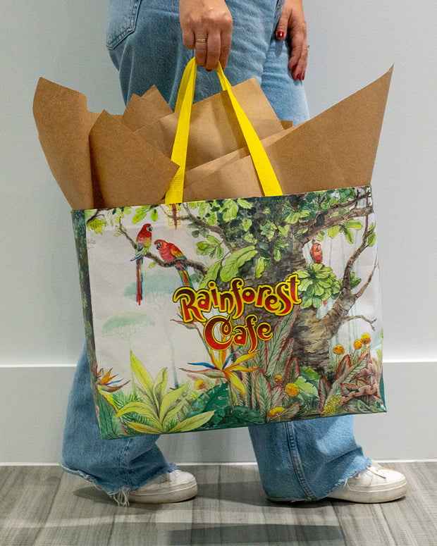 A person is standing holding a colorful shopping bag with the “Rainforest Cafe” logo and illustrations of a tropical rainforest scene, including various birds and lush greenery. The bag appears to be made of a sturdy material and is filled with brown paper, suggesting recent purchases. The person is wearing blue denim jeans and white sneakers, with only the lower half of their body visible in the image. The background is a plain grey wall with a white floor, emphasizing the vivid colors of the shopping bag.