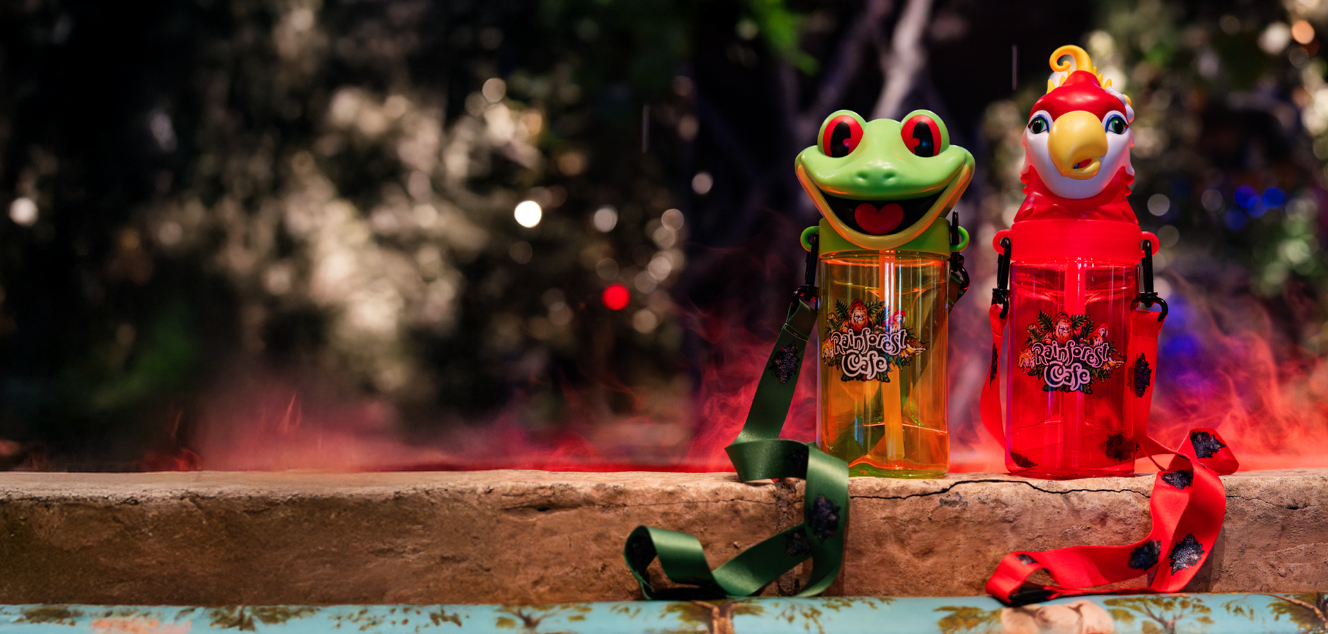 Two colorful souvenir cups resembling a frog and a parrot, adorned with Rainforest Cafe logos, sit on a stone ledge.