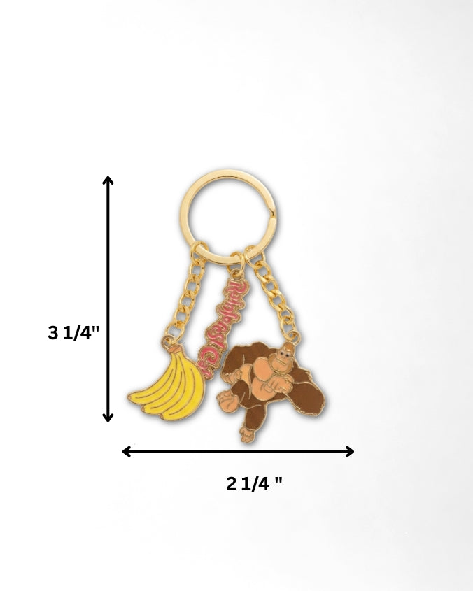 A gold-tone keychain with a banana charm, a monkey charm, and a red script charm, shown with dimension arrows and labels indicating a height of 3 1/4 inches and a width of 2 1/4 inches on a white background.