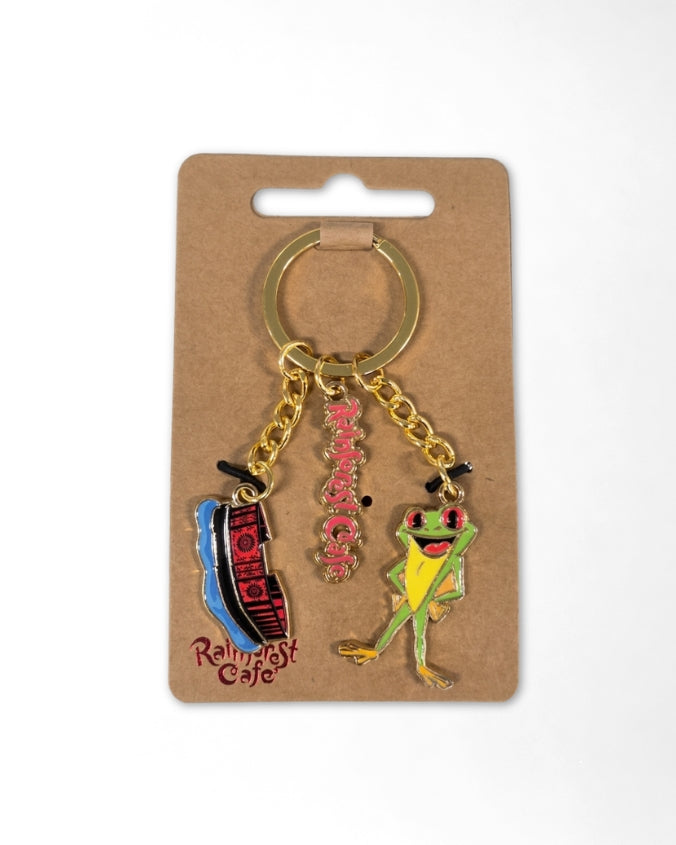 A novelty keychain on a brown cardboard hanger card. The keychain has a gold-tone split ring and chain with three charms: a blue and red striped 'Rolling Stone' magazine cover, a pink rhinestone 'Rolling' text charm, and a green and yellow frog charm wearing a small black top hat.