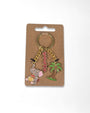 A gold-tone keychain with three charms displayed on a brown cardboard backing. The charms include a silver elephant with pink and yellow details, a pink rhinestone-encrusted word charm, and a green palm tree.