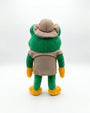A plush toy frog shown from the back, wearing a tan hat, vest, and apron with green arms and legs, and yellow shoes, photographed on a white studio background.