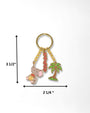 A gold-tone keychain with three dangling charms: a pink elephant holding a yellow pot, a red beaded tassel, and a green palm tree, shown on a white background with measurement arrows and labels indicating 3 1/2 inches tall by 2 1/4 inches wide.