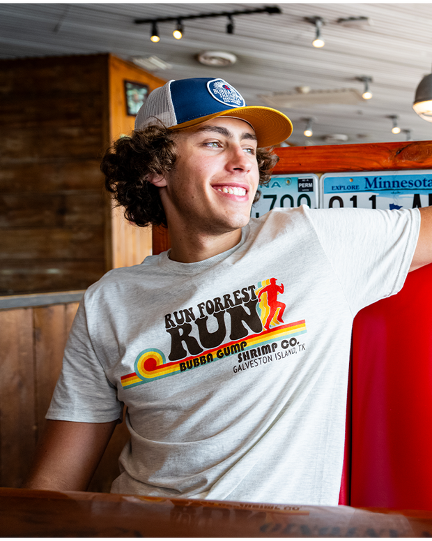 made model sitting in a red booth, looking out. He is wearing a blue and yellow cap with oatmeal shirt that reads in brown letters "run forrest run", "bubba gump shrimp co." under the phrase is lines going straight, with a silhouette of a running person on the right side.