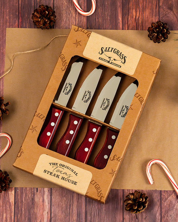 Four steak knives inside a wood box, placed over brown paper that's over a wood surface. Pine cones, candy canes and string frame around the box.