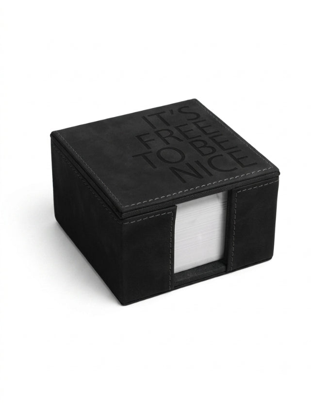 A black square napkin holder with a textured faux-leather finish, featuring the embossed text 'IT'S FREE TO BE NICE' on the lid and a clear window on the front showing white paper napkins inside.