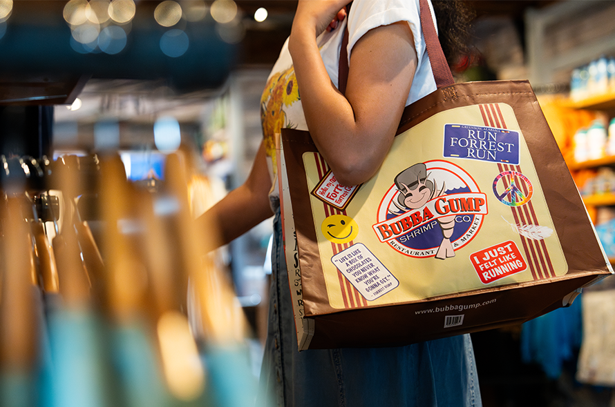 A person holds a Bubba Gump Shrimp Co. tote bag decorated with quotes and graphics, standing in a colorful store.