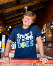 made model resting hands on top of booth edge. he is wearing Navy Scoop Neckshort sleeve tee shirt with blue, white and yellow graphics. The phrase on the shirt says " Stupid is as stupid does". Graphic includes yellow smiley winking face instead "o" letter in the word "does".