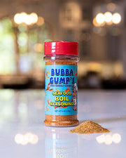 Bubba Gump’s Seafood Boil Seasoning: A clear plastic container with a red cap, containing Bubba Gump’s Seafood Boil Seasoning. The label features colorful graphics and text, including the brand name, ‘CRAB•SHRIMP•CRAWFISH,’ and the net weight (6 oz or 170g). A small pile of the golden-brown seasoning is spilled beside the container.