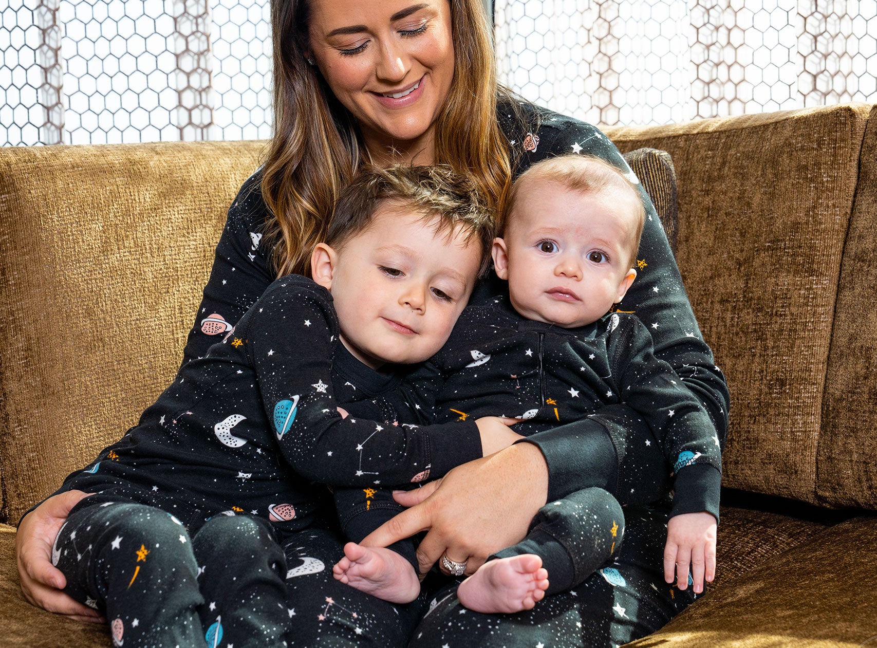 Mom sits on the brown couch with two kids on her lap. All are wearing matching black pajamas with space-inspiring prints. 
