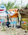Two clear cylindrical water bottles with a blue "AQUARIUM, AN UNDERWATER DINING ADVENTURE", in the center, featuring animal head lids and black lanyards with "AQUARIUM" written on them. Next to them is a green bottle with a T-rex cap and "T-REX" written on the center with a dinosaur image. The background is a gravel surfaced background. 