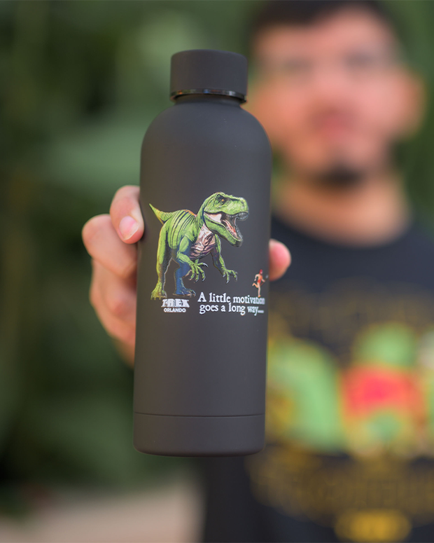 a person holding a black water bottle with a unique and motivational design. The bottle features an illustration of a green Tyrannosaurus rex with the text “T.REX ORLANDO. A little motivation goes a long way.” The background is intentionally blurred to focus on the water bottle, and the person’s face is not visible in the frame. The attire of the person is also not clear due to the close-up shot of the bottle.