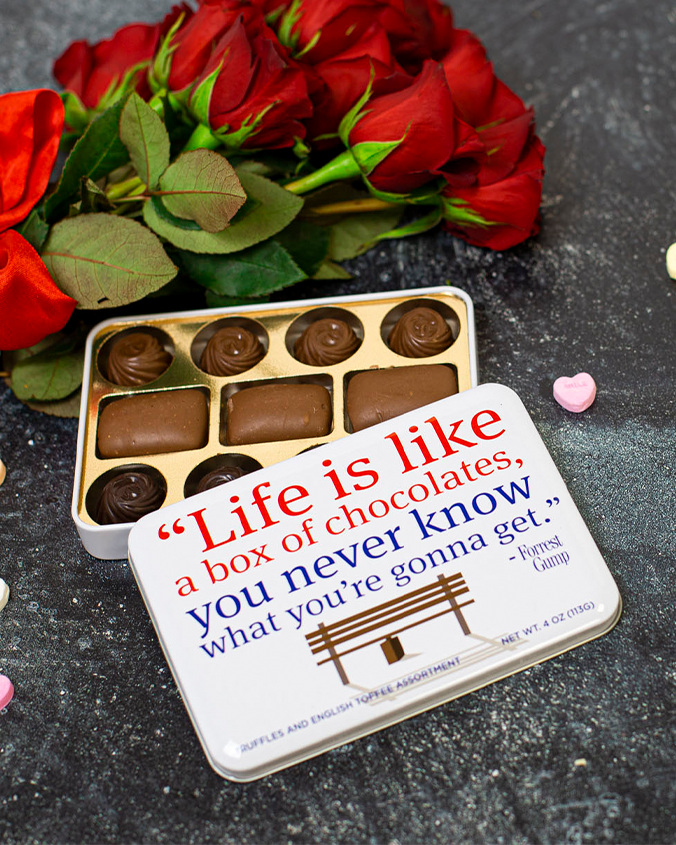 white tin box of chocolates that reads "life is like a box of chocolates, you never know what you're gonna get." - forrest gump. Top of box is removed and laid over the bottom part to show 8 truffles and 3 chocolate squares. next to it is a bouquet of red roses and heart shaped candy.