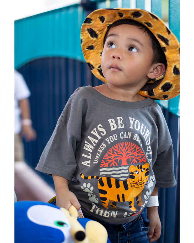 little boy wearing Dark blue/grey kids tee with cartoon tiger design and "Always Be Yourself...Unless You Can Be A Tiger...Then Always Be A Tiger". He is looking up, holding a toy in his hand and wearing an orange bucket hat with black tiger spots.