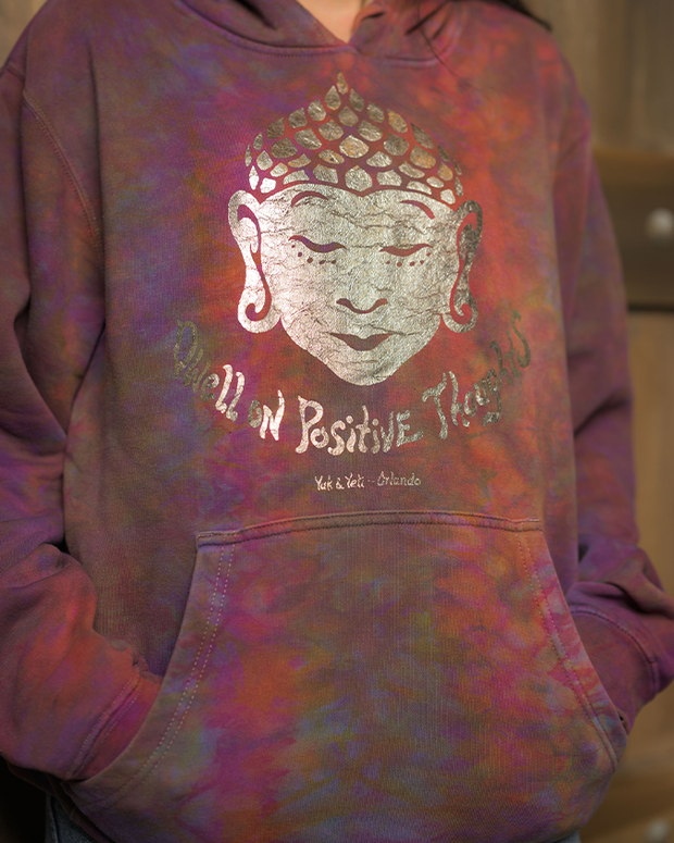 Close up of tie-dye hoodie with a detailed print of Buddha’s face and the phrase ‘Dwell on positive thoughts’ written below. The hoodie’s pattern features shades of purple, pink, and blue, and includes a front pocket. The phrase ‘Yak & Yeti - Orlando’ is also visible in smaller text. The background suggests an indoor setting with a focus on the hoodie.