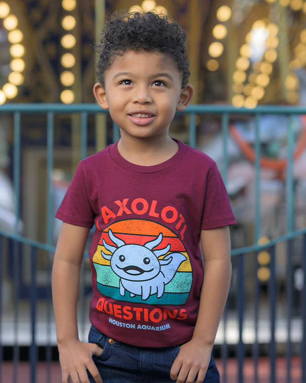 A kid is seen wearing a burgundy t-shirt featuring a colorful and whimsical design with an axolotl at the center, accompanied by the text ‘I AXOLOTL QUESTIONS HOUSTON AQUARIUM’. The background, though blurred, hints at an outdoor evening setting with a fence and illuminated structures, adding a sense of place to the image.