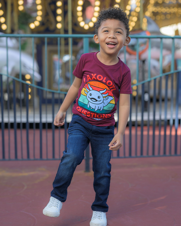 A child is pictured standing in front of a carousel, wearing a vibrant burgundy t-shirt with the playful graphic ‘I AXOLOTL QUESTIONS’. Complementing the t-shirt are blue jeans and white sneakers. The background features the soft glow of carousel lights, adding a festive atmosphere to the amusement park setting.