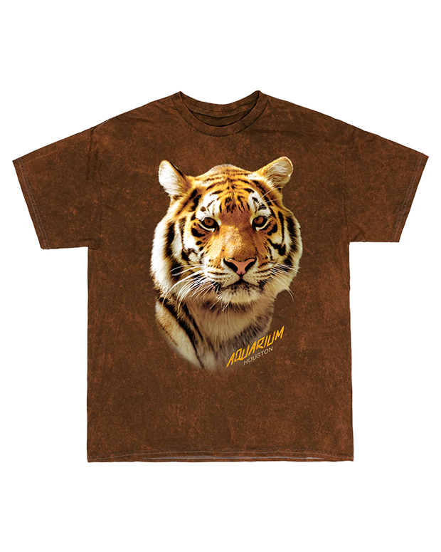A brown t-shirt featuring a large, detailed print of a tiger’s face and the yellow text ‘AQUARIUM HOUSTON’ at the bottom right. The central design on the shirt showcases a realistic and intense image of a tiger’s face, with orange fur and black stripes. The yellow text cleverly identifies the location as AQUARIUM HOUSTON.