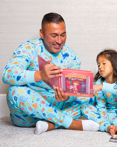 A dad reading a book to his daughter, both wearing matching blue pajamas adorned with colorful under the sea designs, are sitting on the floor.