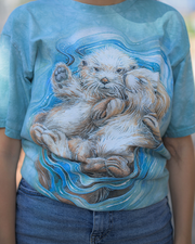 A person is featured wearing a blue t-shirt adorned with a whimsical illustration of two otters. The larger otter seems to be embracing the smaller one, both depicted in a playful and affectionate pose, with their eyes closed as if savoring the moment. They are set against a backdrop of swirling blue and white patterns that evoke the feeling of water currents, highlighting the aquatic theme of the design.