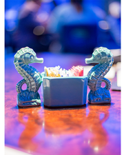 two, blue seahorse shakers faced each other with sugar caddy in center.  They are on a table with the background blurred on people sitting in restaurant. 