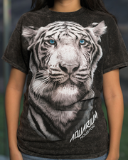 Close up of dark-colored t-shirt featuring a large, detailed print of a white tiger’s face with blue eyes. Below the tiger’s face, there is text that reads ‘AQUARIUM HOUSTON.’ The person stands in front of a blurred outdoor background.