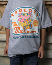 Close up of a kid wearing a grey t-shirt with a dynamic axolotl character design, complete with sunglasses and sneakers, giving an impression of speed and motion. The playful text ‘AXOLOTL TO THE Maxolotl’ surrounds the character in bold, colorful letters, and ‘HOUSTON AQUARIUM’ is noted below. The backdrop includes a portion of a stone wall structure, adding a rustic feel to the image.