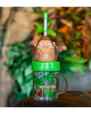 Clear mug with brown handle, iconic tropical Rainforest Cafe design, a green lid, Bamba head, and a clear straw with a green cap. background of stone wall with leaves and palm leaves on it.