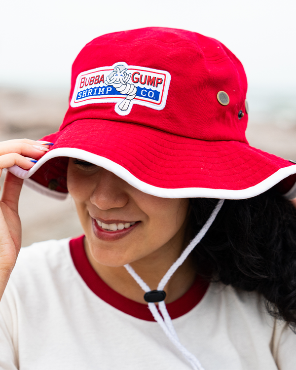 A person holds a red bucket hat with a "Bubba Gump Shrimp Co." logo, wearing a cream shirt with a red collar, against a coastal background.