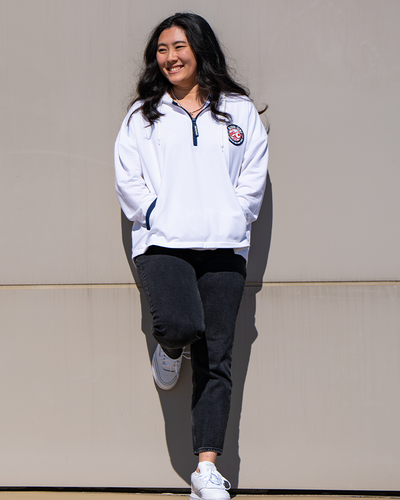 female model with one foot leaned against tan wall. she is wearing white ping pong jacket, black jeans, and white sneakers.