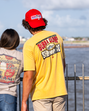 made model standing in front of railing, facing ocean view wearing a red cap and shirt. the shirt is Mustard Yellow Short Sleeve Tee has bubba gump and Co. red, blue, and white charcoal drawing design graphics on the bottom of the drawing small red anchor depicted.