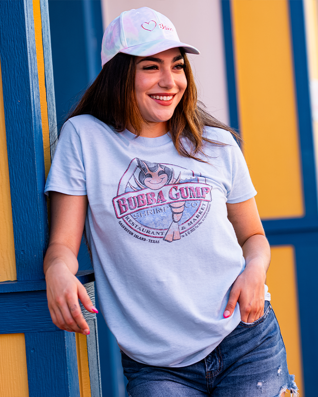 girl leaning on yellow and blue wall wearing Light blue t-shirt with classic distressed Bubba Gump logo and light blue cap.