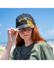 female model wearing Black cap with yellow Yak & Yeti branding and tiger design. she is standing in front of an ocean and holding on to the front of the hat.