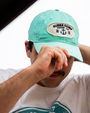 A person wearing a light blue cap with a "Bubba Gump Shrimp Co." patch adjusts their hat outdoors, showcasing a relaxed casual style.