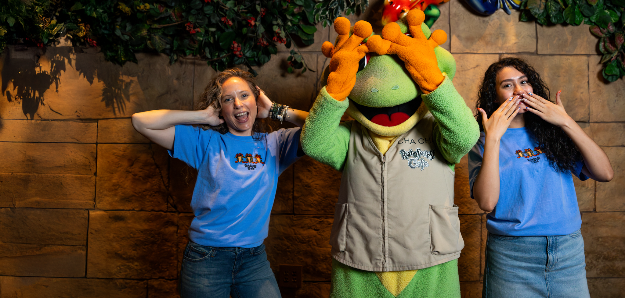 Two smiling individuals in blue shirts pose playfully with a cheerful green frog mascot at a themed restaurant backdrop.