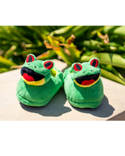 Plush Cha Cha the Frog green slippers with a foam sole placed on stone with palm leaves in back.