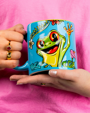 A close-up view of a model in a pink shirts' hands holding a colorful blue mug with a waving frog surrounded by tropical flowers and leaves.