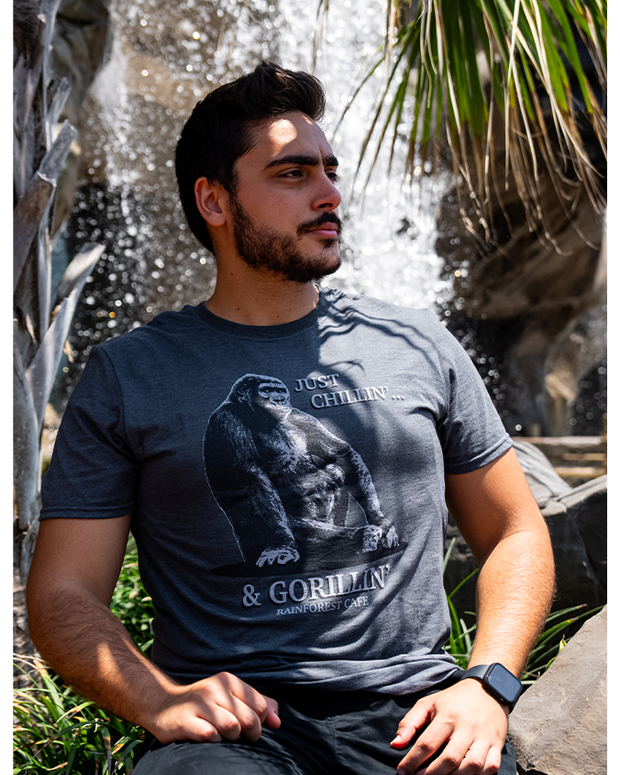 male model sitting in front of a waterfall, under a palm tree. he is wearing navy tee that has Image of a gorilla sitting down. Top left reads "JUst Chillin'..." and bottom of gorilla image reads "& Gorillin'"