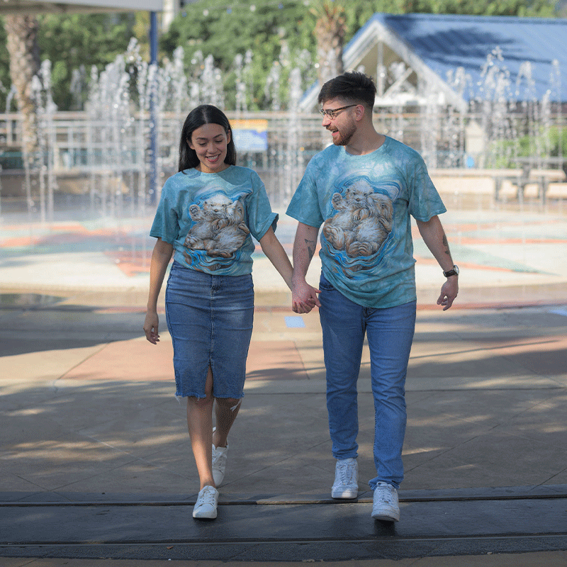 Two individuals walking hand in hand, facing towards a fountain. Both are wearing matching blue tie-dye t-shirts with a large graphic of two otters cuddling on the front. They are in a park-like setting with trees and a clear sky in the background.