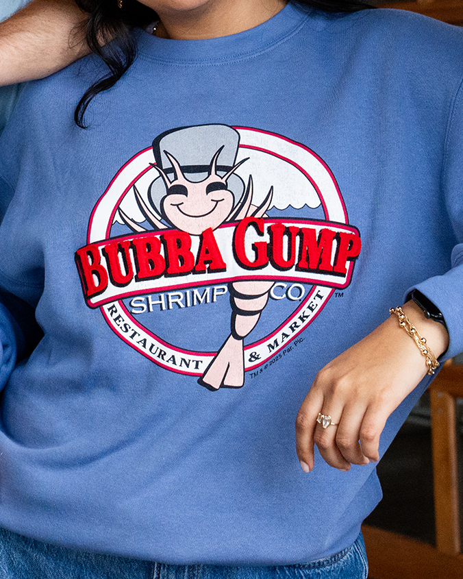 A woman wearing a blue sweatshirt featuring the Bubba Gump Shrimp Co. logo, smiling and showing off the playful design.