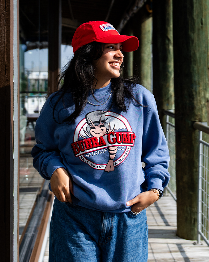 A person wearing a blue sweatshirt featuring the Bubba Gump Shrimp Co. logo, paired with denim jeans and a red cap, stands on a walkway.