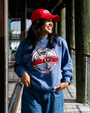 A person wearing a blue sweatshirt featuring the Bubba Gump Shrimp Co. logo, paired with denim jeans and a red cap, stands on a walkway.