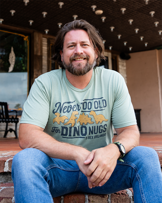 A man sits casually on steps, wearing a light green t-shirt featuring a playful dinosaur nugget graphic and blue jeans.