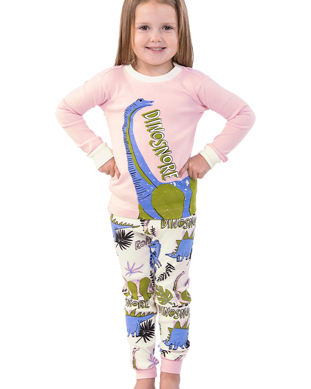 A kid in a cozy pink long-sleeve shirt adorned with a colorful dinosaur illustration and the word ‘DINOSNORE’, paired with pajama pants featuring a playful dinosaur pattern, against a plain white background.