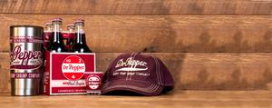 dr pepper 4 count bottles, silver tumbler, shot glass and burgundy cap placed together on left side. background is a tan wood.