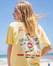 back view of female model standing in front of ocean view wearing Light yellow shirt with a red and green dragon in the middle and words "Power, Strength, Honor" in Chinese and English.