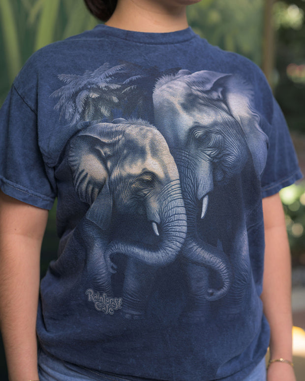 A blue t-shirt featuring a graphic print of two elephants with their trunks intertwined, set against the shirt’s darker blue fabric. Below the elephant design, the text ‘Rainforest Cafe’ is prominently displayed.