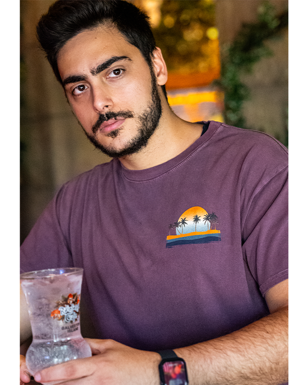 male model wearing brown shirt with sunset of palm trees on beach on left chest. he is holding a rainforest cafe logoed glass cup.