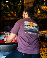 male model at bar inside rainforest cafe. he is wearing brown shirt that has a framed image of a sunset with palm tress on beach. under image reads "rainforest cafe".